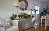White-painted wicker armchair next to festive arrangement and table lamps on sideboard with Christmas tree in background