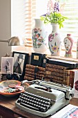 Vintage typewriter and ceramic bowl in front of stacked antiquarian books on table below window with vases on sill