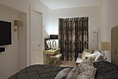 Elegantes Doppelbett mit dunkelbrauner Tagesdecke und drapierten Kissen, Schminktisch vor Fenstertür mit geschlossenem Vorhang