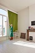 Woman standing by window with green curtain in period apartment with white wooden floor