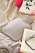 Hot-water bottle with pale grey knitted cover, blanket and book on bed