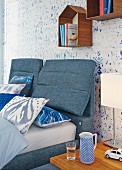 Double bed and headboard upholstered in denim below small, house-shaped shelving modules on wallpaper with blurry pattern