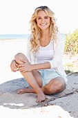 A young blonde woman on a beach wearing a white top, jacket and shorts