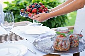 Glass dish of fresh berries held in hands and preserving jars on tray on garden table