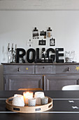 Tealights and candles on table in front of decorative letters on grey sideboard and collage of postcards on wall