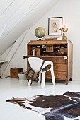 Cowhide rug on white floor and white shell chair at antique wooden bureau with globe on top in attic room