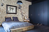 Doppelbett mit graublauer Bettwäsche vor Natursteinwand, seitlich dunkelblauer Kleiderschrank