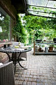 Garden table with metal base and mosaic top on roofed terrace with cobbled floor and outdoor kitchen in background