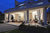Villa in Nachtstimmung, Blick auf beleuchtete Veranda mit gerafften Vorhängen und Sitzmöbeln