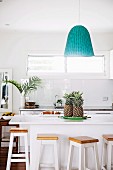 Wooden bar stool around white kitchen counter with pineapple, pendant lamp above with turquoise lampshade