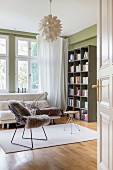 Sheepskin blanket on classic chair, stool and sofa below window next to bookcase against green wall