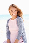 A young blonde woman on a beach wearing a pastel pink dress and a cardigan