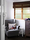 Blick durch offene Zimmertür auf grauen Lesesessel mit Stofftieren und Kissen, neben Sprossenfenster mit Bambusrollo im Kinderzimmer