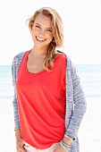 A blonde woman on a beach wearing a red top and a cardigan