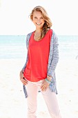 A blonde woman on a beach wearing a red top, pink trousers and a cardigan