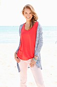 Blonde Frau in rotem Shirt, rosa Hose und Strickjacke am Strand