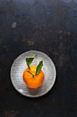 A clementine with leaves on a plate on a dark surface