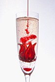 Red syrup being added to a hibiscus flower in a glass of champagne