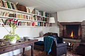 Antique console table below wall-mounted bookshelves and armchair in front of open fire