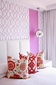 Three colourful scatter cushions on bed against wall covered in purple-patterned wallpaper