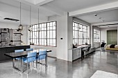 Concrete floor, factory windows, dining table and blue chairs in industrial loft apartment in shades of grey