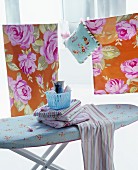 Striped cloths on ironing board in front of rose-patterned bed linen hung on washing line