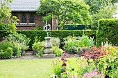 Sommerlicher Garten mit blühenden Stauden und Steinstele mit Sonnenuhr vor Hecke und Ziegelfassade