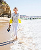 A woman wearing a cashmere cardigan, a short sleeved patterned blouse and white trousers walking along a beach