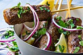 Cevapcici on wooden skewers with onions, parsley and lemon in a grey bowl