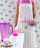 Woman wearing striped apron standing in front of floral wallpaper holding bowl of strawberries