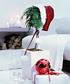 Small conifer wearing Father-Christmas hat next to red bauble with white lettering on top of wrapped gift