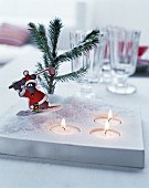 Weihnachtliche Cartoon-Figur auf Teelichthalter mit Kunstschnee, Tannenzweig und brennenden, eingelassenen Teelichtern