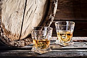 Zwei Gläser Whisky mit Eiswürfeln vor altem Holzfass