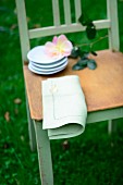 Stack of small plates, cut rose and linen napkin on chair seat