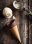 A melting scoop of vanilla ice cream, a chocolate-dipped cone and an old ice cream scoop on a wooden surface