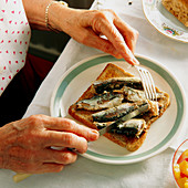 Elderly woman's hands use cutlery to eat sardines