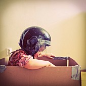 A little girl wearing a motorbike helmet sitting in a cardboard box