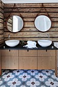 Custom washstand with twin white washbasins below round mirrors hung on rustic log-cabin wall