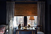 Table lamp on antique desk in front of window with bamboo roller blind and matching blue and white patterned curtains, armchair and chair seat