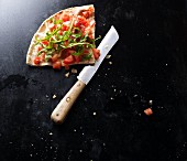 A slice of bruschetta pizza with a knife on a black baking tray