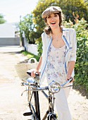 A woman pushing a bicycle wearing a striped blouse, a floral print top, white trousers and a flat cap