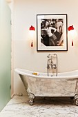 Freistehende, versilberte Badewanne mit nostalgischer Wandarmatur unter roten Wandleuchten und femininer Fotografie