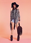 A woman wearing a grey, coarse-knit ponco, denim shorts, fringed boots ad a floppy hat
