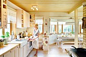 Pale wooden ceiling, woman wearing apron in front of kitchen counter and dining area in window bay in open-plan kitchen