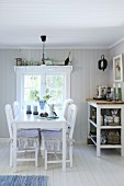 White dining table and chairs next to shelves of crockery below window in Swedish house