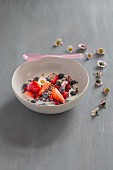Beeren mit Joghurt in weisser Porzellanschüssel mit Kakaonibs, rosa Löffel und Gänseblümchen