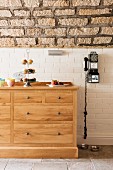 Black vintage payphone on white brick wall next to solid oak chest of drawers below vaulted stone ceiling