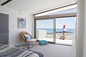 Airy bedroom with window front and sea view, woman on glass balustrade on terrace