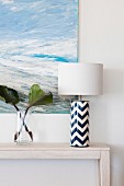 Elegant, blue and white table lamp next to a glass vase in front of a maritime painting