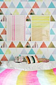 Two frame graphic artworks on colourful wallpaper above scatter cushions on bed